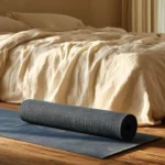A cream-linen bed and folded blanket beside a blue yoga mat partly rolled on a wooden floor.