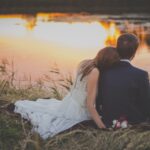 sitting woman leaning on man's shoulder facing lake during golden hour