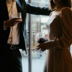 A sophisticated couple in formal attire holding drinks in a moody restaurant atmosphere.